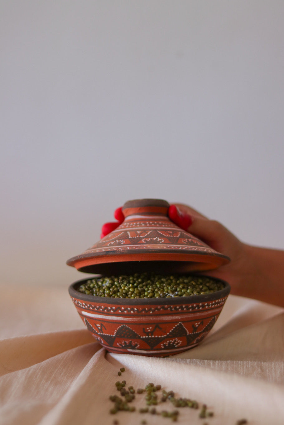 a red hand painted pottery serving bowl with hand painted lid is sitting on top of a white cloth with is lid being open by a woman