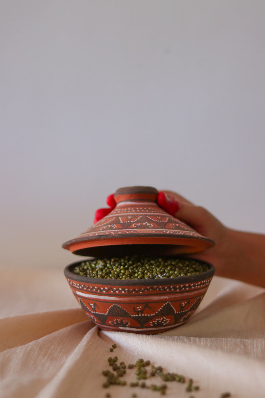 a red hand painted pottery serving bowl with hand painted lid is  sitting on top of a white cloth with is lid being open by a woman