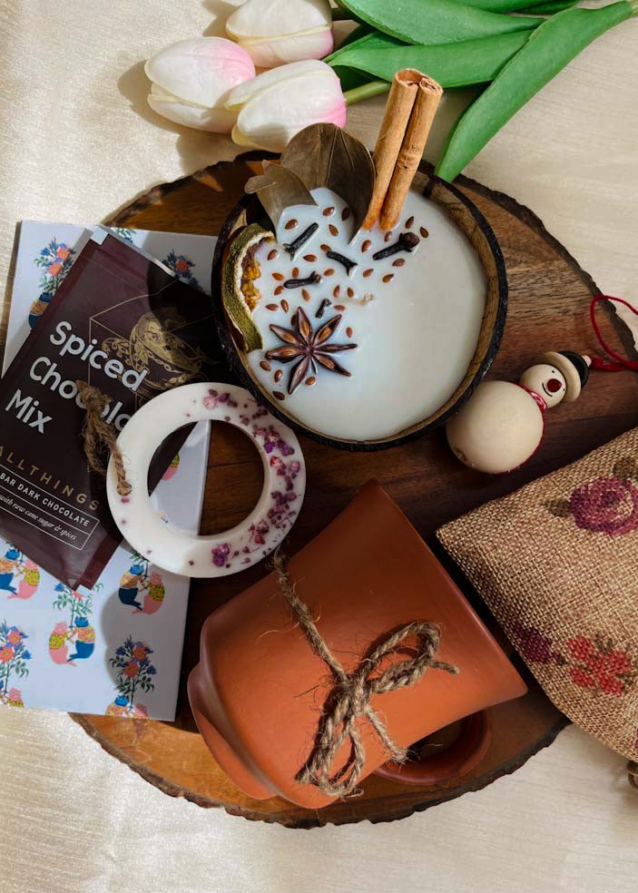 hand poured coconut candle decorated with spices, hand poured wax tag decorated with flowers, a coffee mug and jute bag along with dark chocolate packet is kept on a table