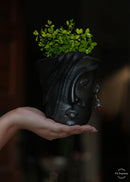 A black clay planter in a shape of a woman's face plabted with green plants is being held in a woman's hand