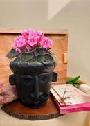 a black planter in a shape of a man's face, decorated with pink flower  is kept on a table along with chocolate and cards kept. and akkaara wooden box in the background