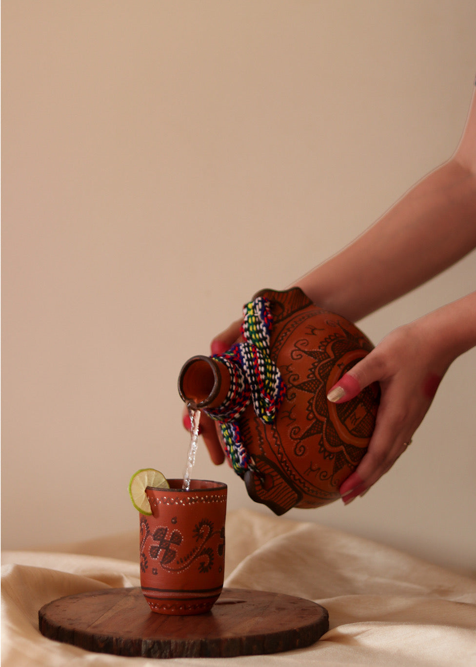 A red hand painted clay water bottle pouring water in a clay hand painted glass