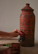 A red hand painted clay water dispenser kept on the table while dispensing water in a hand painted red clay glass