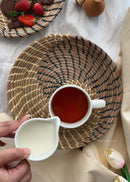 hand woven sikki plate with swirl pattern in blue is kept on the table, a tea cup is kept on it and a woman is pouring milk in the cup