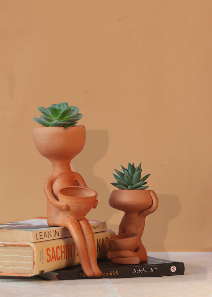Boy shaped clay planters with green plants are kept on a pile of book