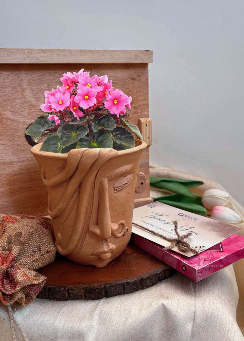 A woman shaped white planter is decorated with pink flower and is kept on the table along with chocolates and the akkaara gicting box