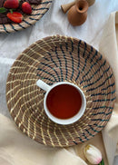 hand woven sikki plate with swirl pattern in blue is kept on the table, a tea cup is kept on it.