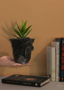 A black clay planter in a shape of a woman's face plabted with green plants is being held in a woman's hand and books are kept in the background