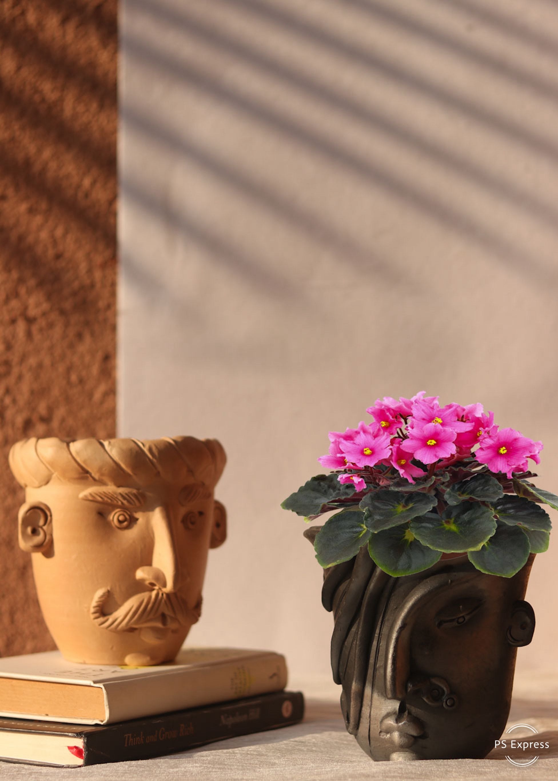 A black clay planter in a shape of a woman's face with pink flower is a kept on the table. behind it is a clay white planter in a shape of man's face kept on the book pile