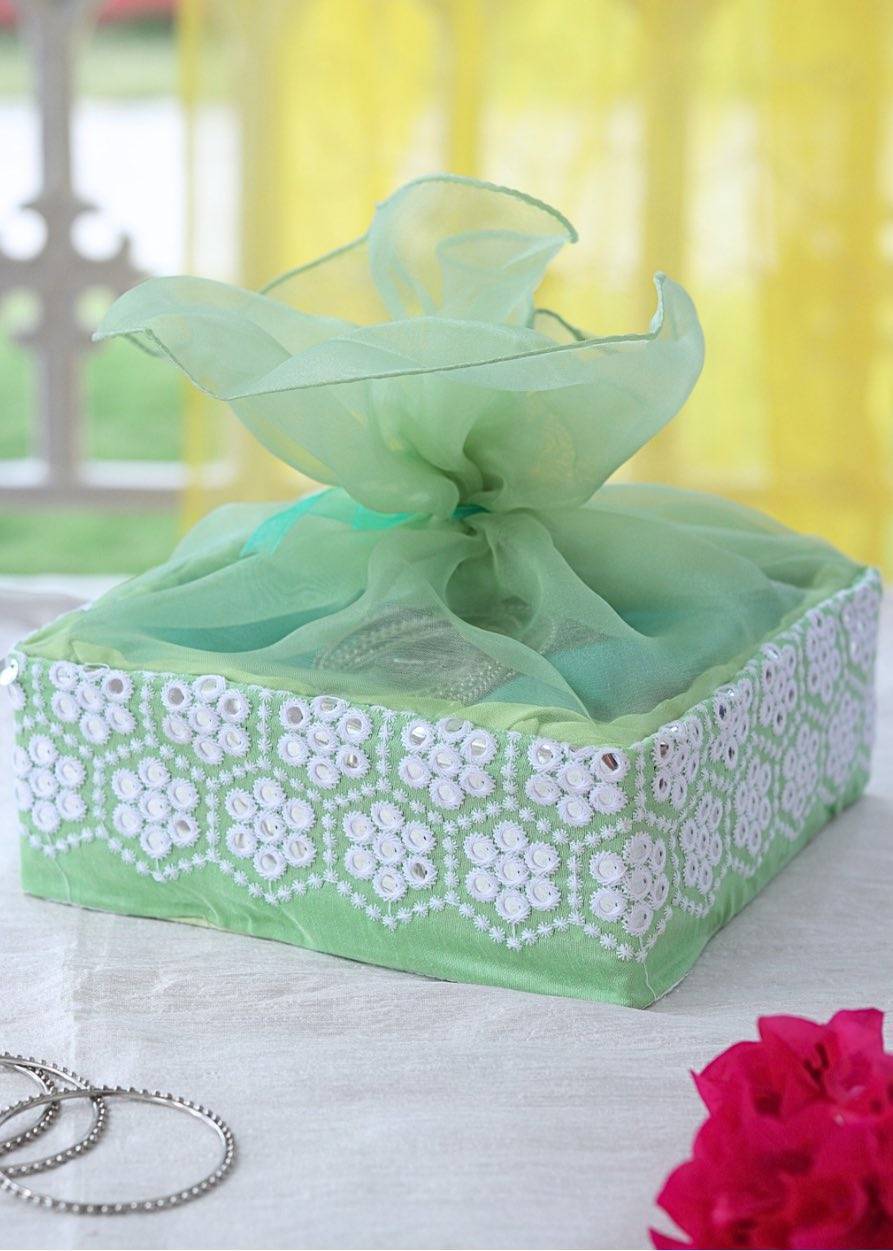 A pastel green embroidered gift box with a decorative green ribbon on top, placed on a table with a yellow background and a floral pattern in the background.