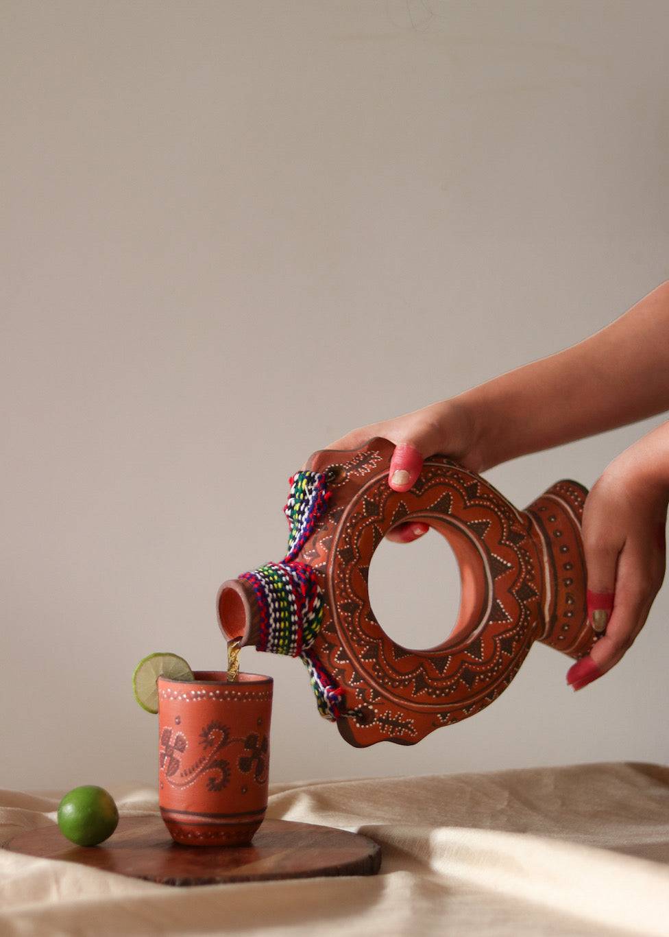 A red hand painted clay water bottle pouring water in a clay hand painted glass